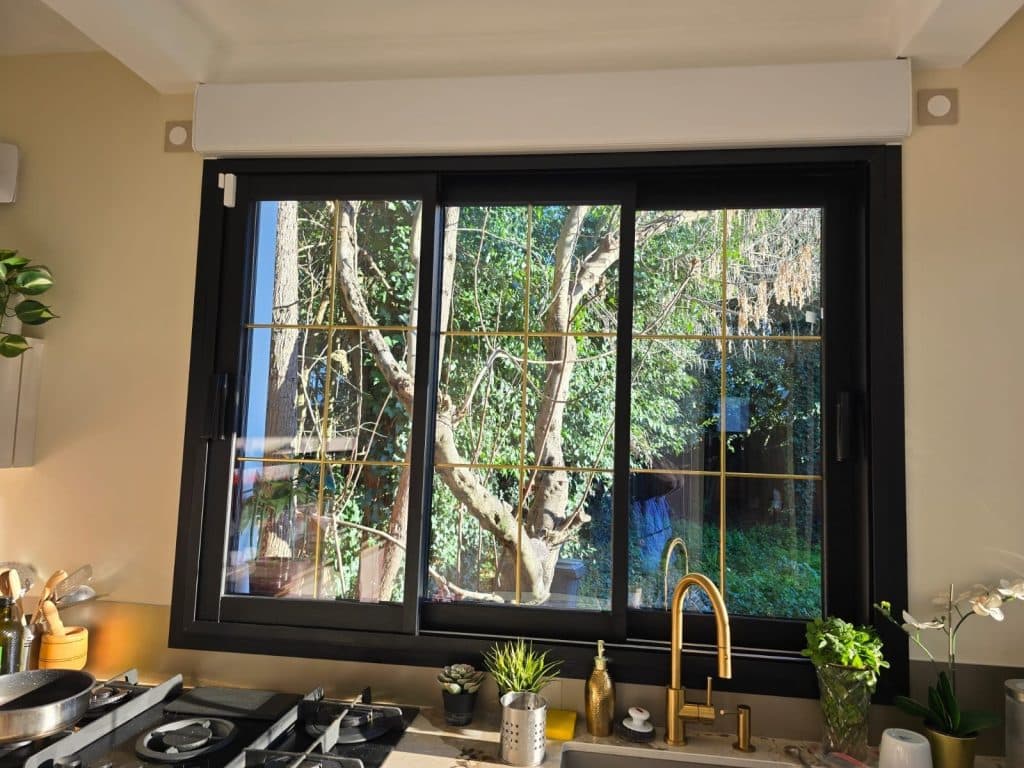 Fenêtre de cuisine en aluminium noir avec petits-bois laiton, robinet doré et vue sur le jardin arboré